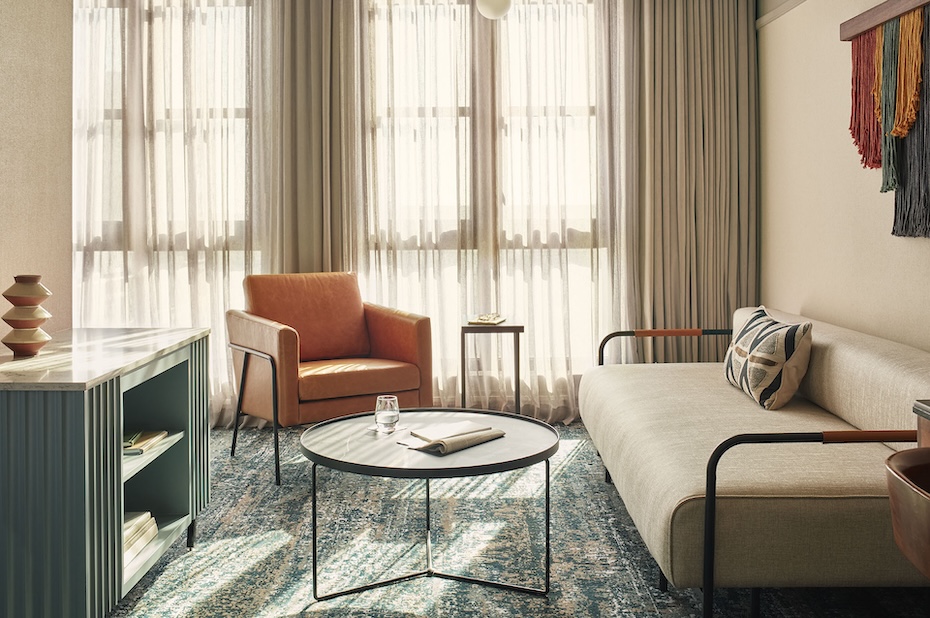 A cozy lounge area within a hotel suite featuring a low-profile cream sofa and an cognac leather armchair. A round black coffee table sits on a textured blue and grey patterned carpet, with soft sunlight filtering through large windows.