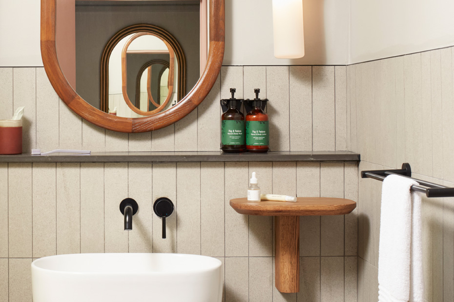 Close-up of a hotel vanity area featuring an oval mirror, black wall-mounted faucets, and a small wooden wall-mounted shelf. Two green bottles of premium hand and body wash are neatly displayed on a stone ledge.